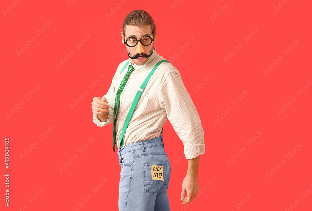 Sticky note with text KICK ME on buttocks of young man with funny disguise against color background. April Fools Day celebration Sticky note with text KICK ME on buttocks of young man with funny disguise against color background. April Fools Day celebration