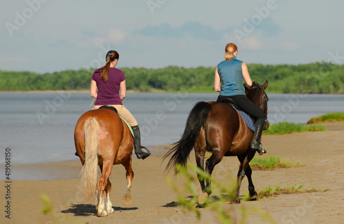 Two women are riding on horseback on beach, back view.