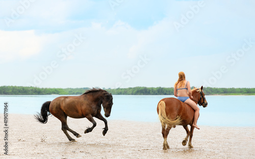 Caucasian woman in bathing suit is riding on horseback on beach and her another free horse is running and is playing. Female rider and two her mare are outdoors.