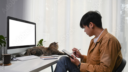 Side view of young man graphic designer working with digital tablet and cute cat lying in front of him. photo
