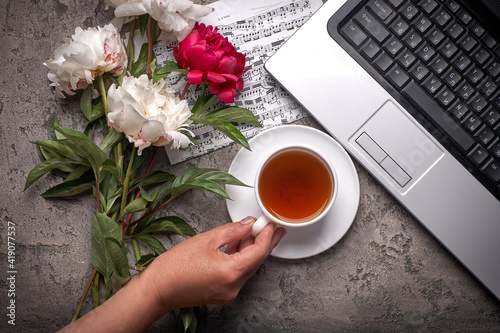 Coffe, peonies and laptop on grey vintage background. Women hand holds cup of coffee.