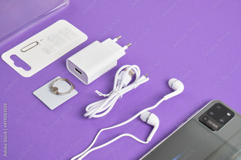 a set of accessories for a smartphone, phone, charger, headphones, a ring-holder, adapters for SIM cards on a purple background a set of accessories for a smartphone, phone, charger, headphones, a ring-holder, adapters for SIM cards on a purple background