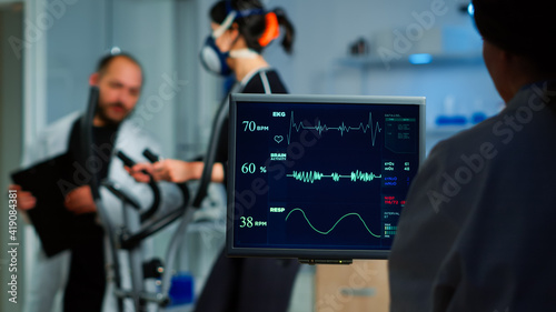 Fototapeta Naklejka Na Ścianę i Meble -  Medical researcher examining EKG image showing on monitor while patinet with mask running on cross trainer testing heart rate using electrodes.Doctor monitoring physical endurance in science sport lab