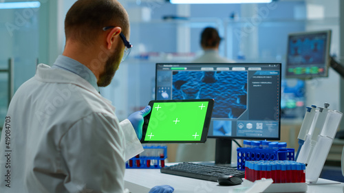 Researcher holding and looking at tablet with chroma key display in modern equipped lab. Team of microbiologists doing vaccine research writing on device with green screen, isolated, mockup display.
