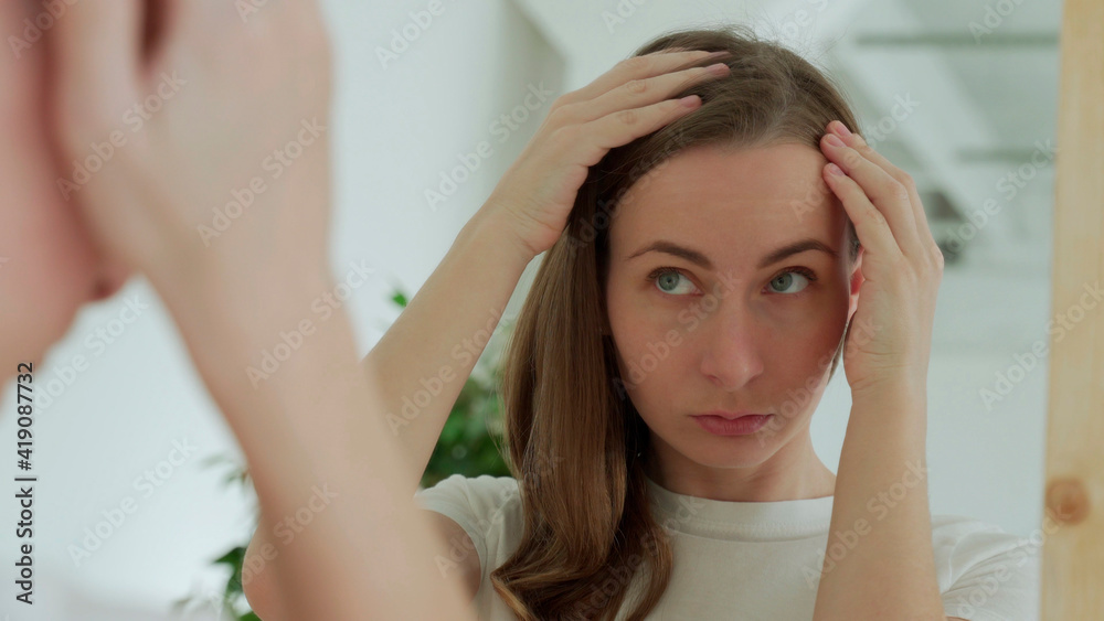 Sad upset woman standing by mirror looking at matted fragile strand in her hand thinking about natural medicinal shampoo restoring hair structure Sad upset woman standing by mirror looking at matted fragile strand in her hand thinking about natural medicinal shampoo restoring hair structure