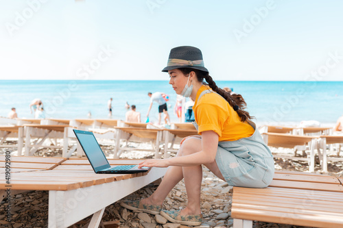 Freelance. A woman with a medical mask on her face, sitting on a chaise longue and typing on a laptop. In the background, the beach and the sea. The concept of remote work during a pandemic