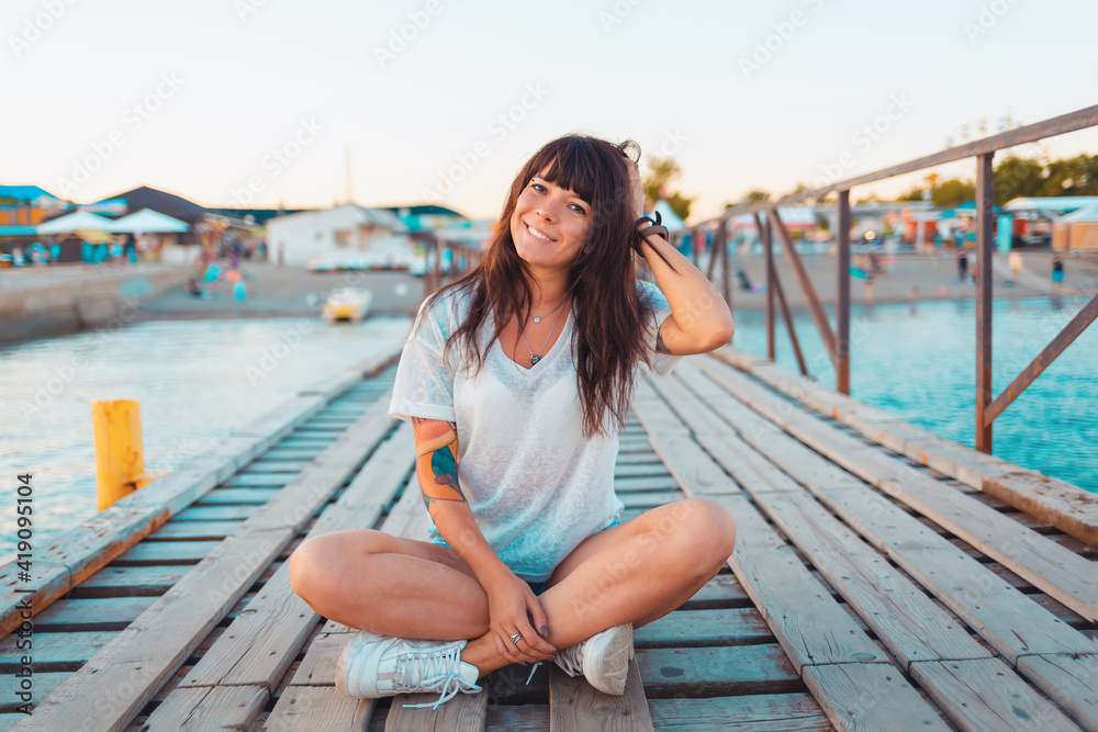 A pretty woman with a tattoo on her arm, smiling and sitting cross-legged on a dock by the sea. Concept of summer vacation