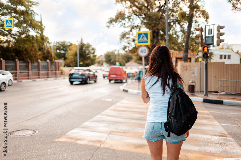 Summertime. Woman with a tattoo on her arm with backpack, standing at a pedestrian crossing. Rear view. Concept of traffic and safety rules