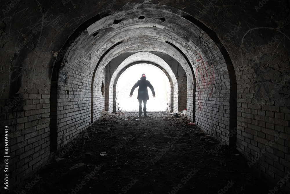 Terrible silhouette of a man with two burning lights on his head, with outstretched arms at the entrance to an old abandoned brick underground tunnel. 
