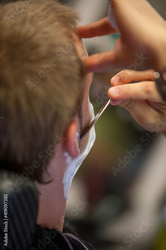 Young bearded man getting haircut by professional at modern barbershop. Beard and sideburns shaving and shaping with vintage straight razor. Barber shop advertising concept. . High quality photo