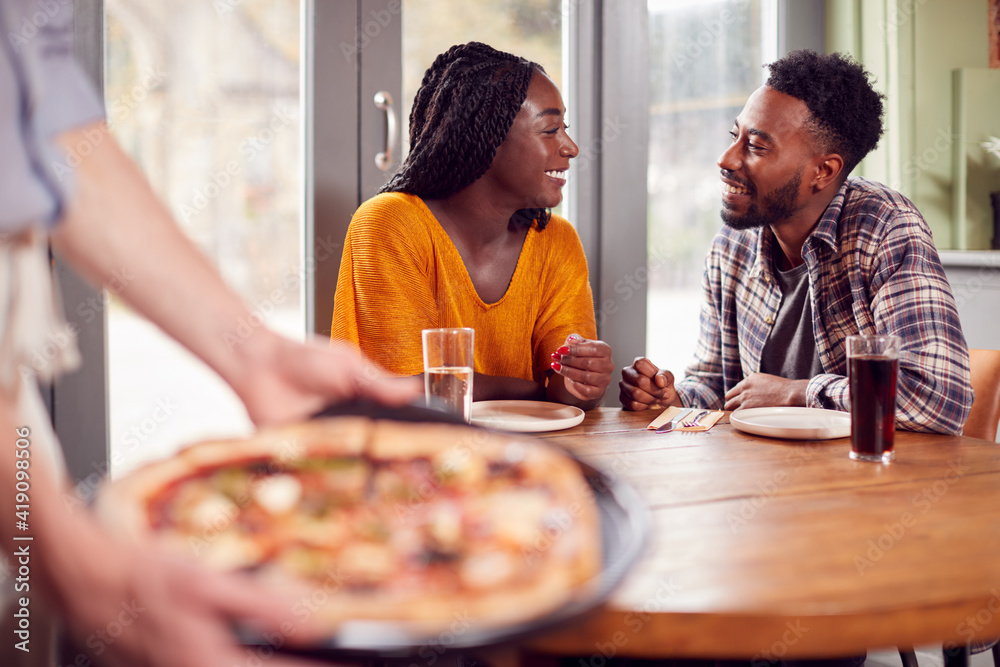 Smiling Couple On Date In Restaurant Together With Waiter Holding Pizza In Foreground Smiling Couple On Date In Restaurant Together With Waiter Holding Pizza In Foreground
