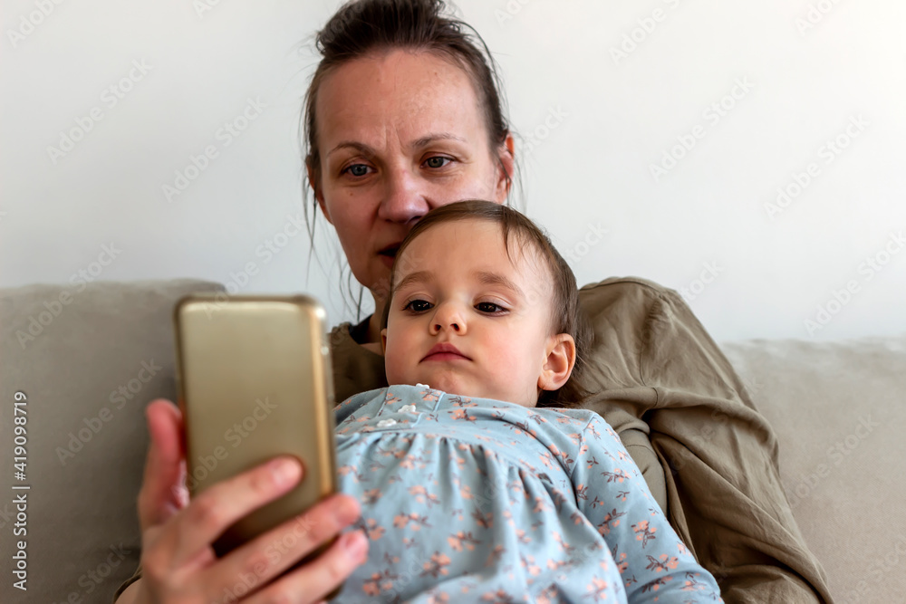 Beautiful young mother at home with little baby girl in her lap, sitting in the living room, holding smartphone. Single mum and child browsing mobile device app, watching cartoon, sitting on cozy sofa Beautiful young mother at home with little baby girl in her lap, sitting in the living room, holding smartphone. Single mum and child browsing mobile device app, watching cartoon, sitting on cozy sofa
