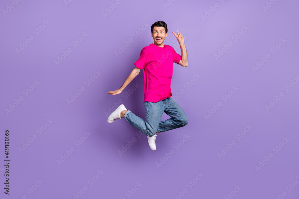 Full size photo of young happy cheerful excited smiling man jumping showing v-sign isolated on purple color background