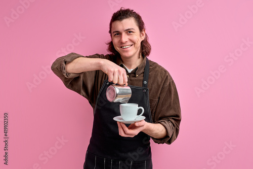 male waiter pouring delicious coffee into cup, handsome guy recommending you to taste this sort of coffee wearing apron isolated over pink vivid color background