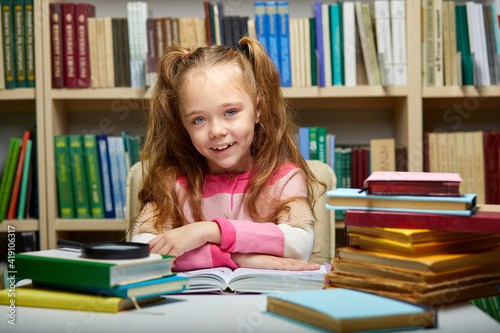 kid girl sits with books at table in library,child in bookstore, surrounded by colorful books for school, she looks at camera smiling