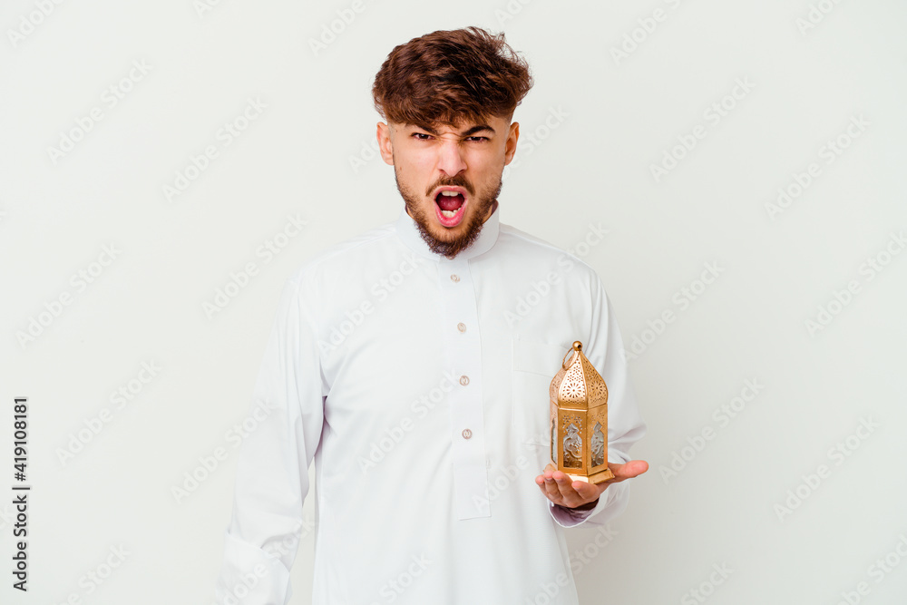 Young Moroccan man wearing a typical arab clothes holding a ramadan lamp isolated on white background screaming very angry and aggressive.