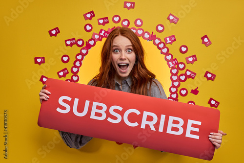 Redhead female asking recommending to subscribe, pay attention, holding title in hands. Social media concept. Isolated yellow background