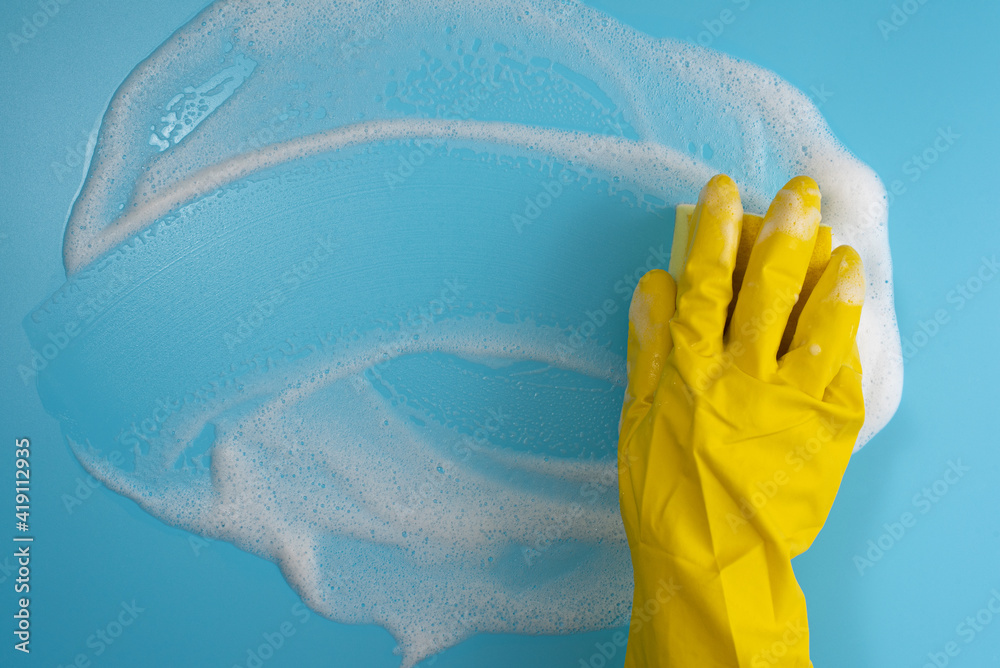 Female hand in protective yellow glove, sponge and foam on blue background. House cleaning concept. Top view. Copy space.