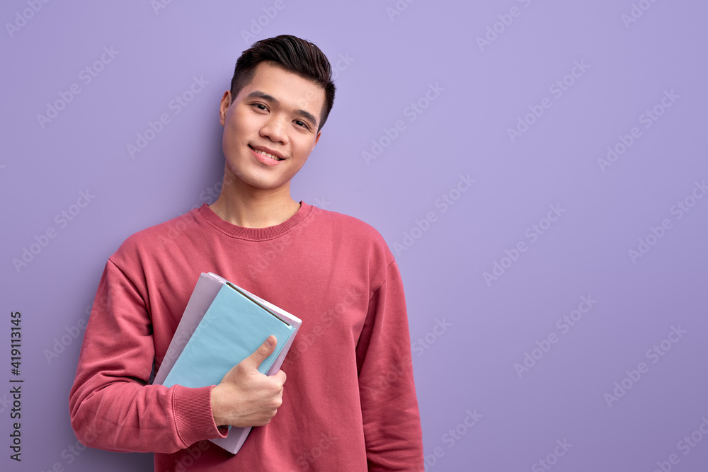 Pleasant Asian student with book in hands enjoy education and university, posing at camera, wearing red casual shirt, isolated on purple studio background Pleasant Asian student with book in hands enjoy education and university, posing at camera, wearing red casual shirt, isolated on purple studio background
