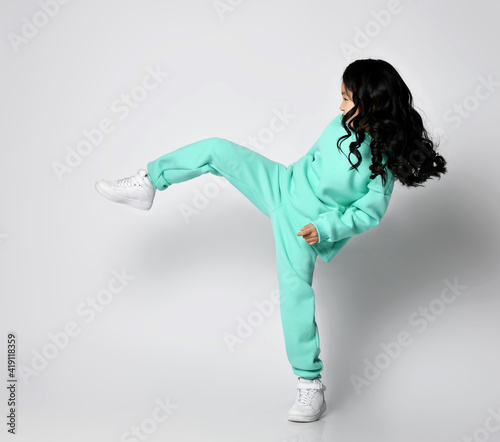Frolic playful asian kid girl in pastel green, mint color sports suit hoodie and pants stands sideways kicking, playing football, soccer, making big step over white background
