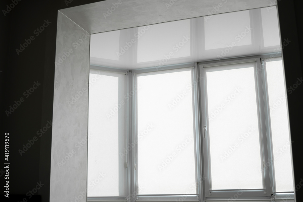 White fabric roller blinds on the plastic window on a balcony in the living room with a reflection in a stretch ceiling. White fabric roller blinds on the plastic window on a balcony in the living room with a reflection in a stretch ceiling.