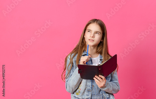  pensive white girl 10 years old in a blue denim jacket with a notebook and pen on a pink background