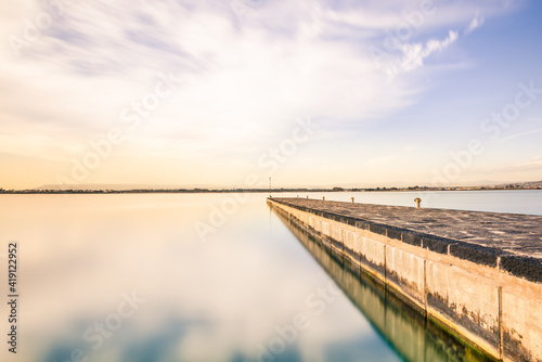 Long exposure of a bridge at evening before sunset. Blue sky, white clouds, sky mirrored in water, calm ocean