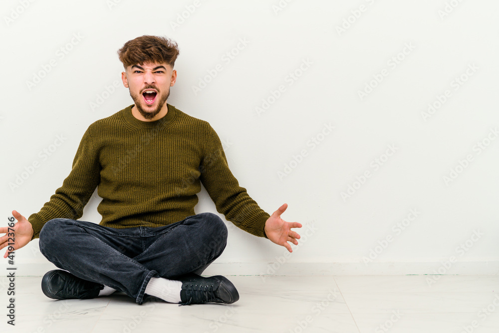 Young Moroccan man sitting on the floor isolated on white background shouting very angry, rage concept, frustrated.