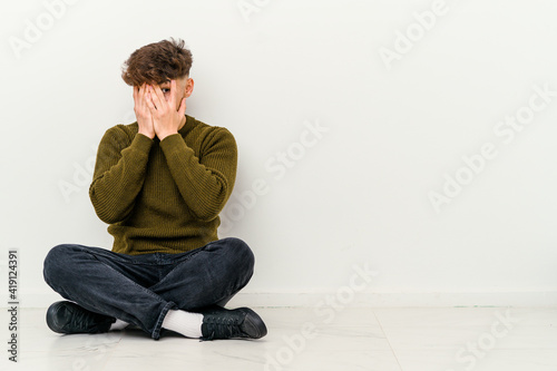 Young Moroccan man sitting on the floor isolated on white background blink through fingers frightened and nervous. © Asier