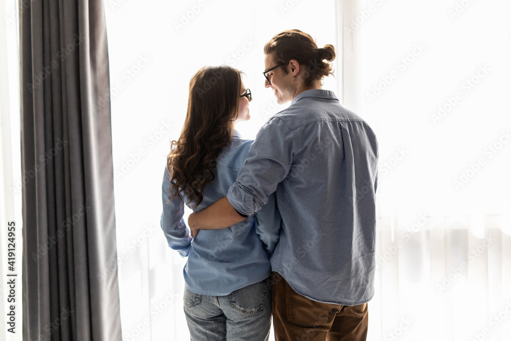 Back view of young couple looking out the window and hugging while having a breakfast at home Back view of young couple looking out the window and hugging while having a breakfast at home