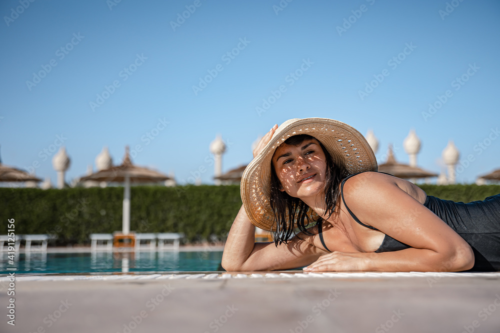 A girl in a straw hat lies sunbathing near the pool on a sunny day.