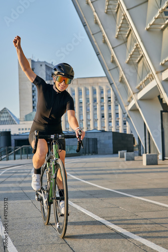 Excited professional female cyclist in black cycling garment and protective gear smiling, raising her arm, looking satisfied with the result of her training