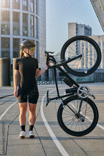 Full length shot of cheerful professional female cyclist in black cycling garment and protective gear looking aside, posing with her bicycle standing vertically outdoors on a sunny day