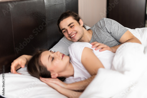 Happy handsome young man lying and waking his woman in bed. Couple wake up together.
