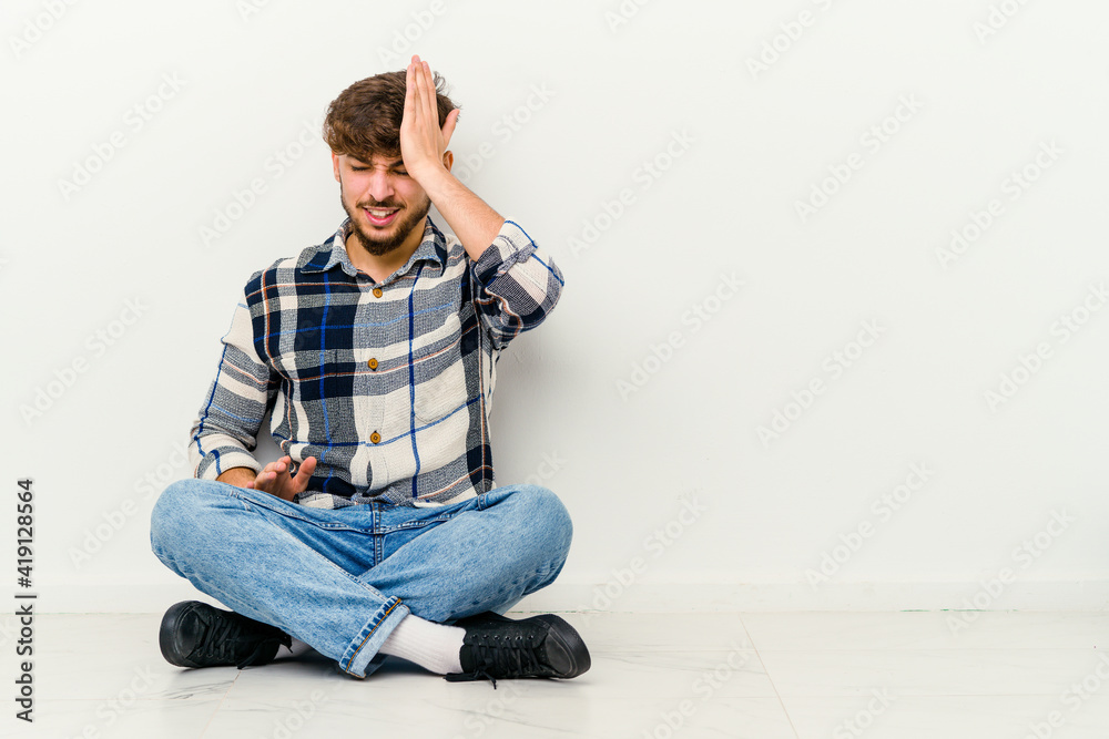 Young Moroccan man sitting on the floor isolated on white background forgetting something, slapping forehead with palm and closing eyes. Young Moroccan man sitting on the floor isolated on white background forgetting something, slapping forehead with palm and closing eyes.