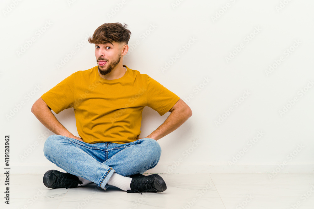 Young Moroccan man sitting on the floor isolated on white background funny and friendly sticking out tongue. Young Moroccan man sitting on the floor isolated on white background funny and friendly sticking out tongue.