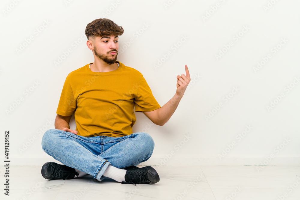 Young Moroccan man sitting on the floor isolated on white background pointing with finger at you as if inviting come closer. Young Moroccan man sitting on the floor isolated on white background pointing with finger at you as if inviting come closer.