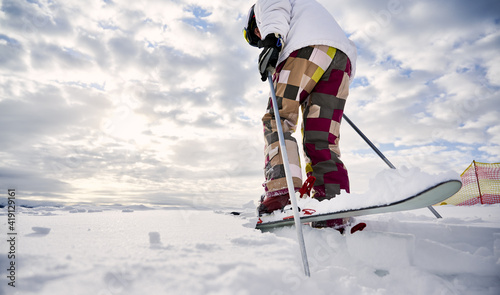 Close-up, low angle view snapshot of skier's legs wearing colorful ski pants and white jacket, making step forward on white snowy surface, ski tour. Copy space. Concept of winter sport activities.