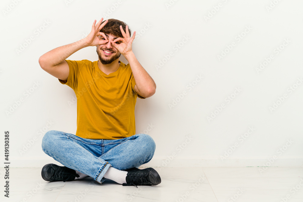 Young Moroccan man sitting on the floor isolated on white background showing okay sign over eyes Young Moroccan man sitting on the floor isolated on white background showing okay sign over eyes