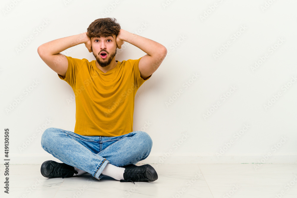 Young Moroccan man sitting on the floor isolated on white background covering ears with hands trying not to hear too loud sound.