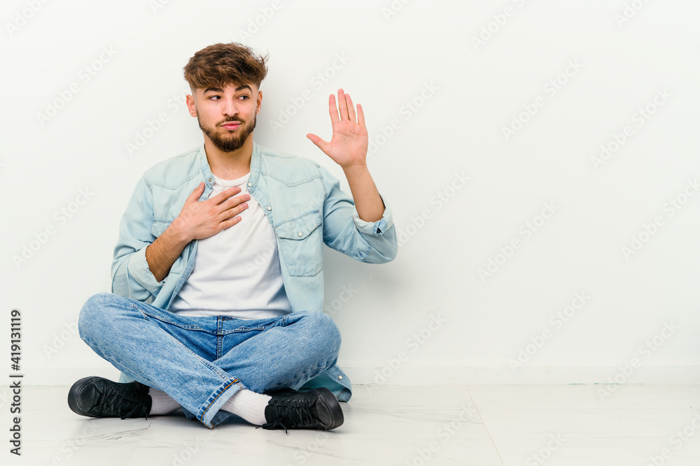 Young Moroccan man sitting on the floor isolated on white background taking an oath, putting hand on chest. Young Moroccan man sitting on the floor isolated on white background taking an oath, putting hand on chest.