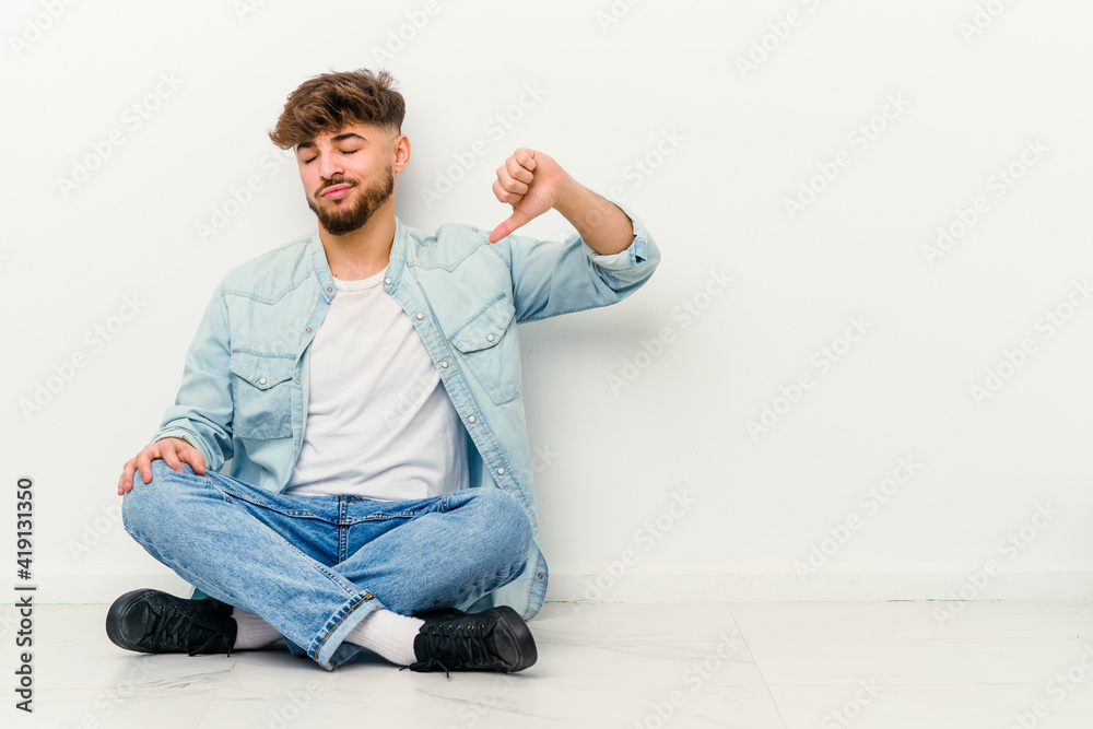 Young Moroccan man sitting on the floor isolated on white background showing a dislike gesture, thumbs down. Disagreement concept.