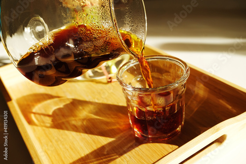 Freshly brewed black coffee is poured into a glass cup on a bamboo tray in sunlight