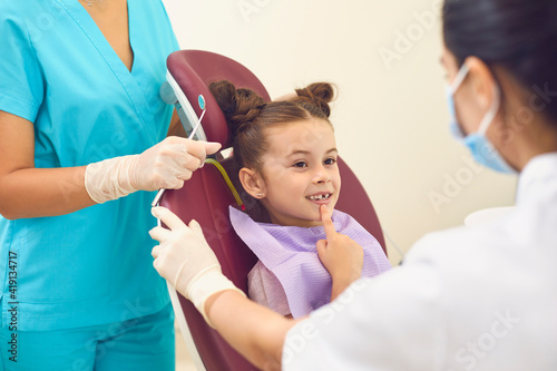 Little girl in a dental office shows two female dentists a tooth that worries her.