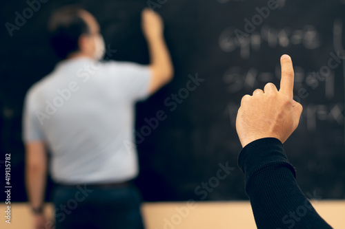 Student raising hand with the teacher wearing face mask writing on the blackboard. Covid situation. New normal. Pandemic.