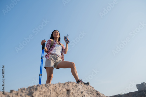 fit young woman hiking in the mountains standing on rocky summit ridge with backpack and pole looking out over landscape. happy female taking a break on a hike. woman drinking water while out hiking