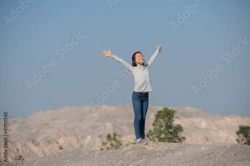 Happy Little asian girl child hiking in the mountains standing on a rocky summit ridge and pole looking out over landscape. hiker relaxing on top of a mountain and enjoying valley view.