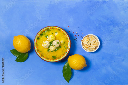 A bowl of avgolemono chicken soup, with meatballs. On a bright blue background with lemons, crackers and peppers. Top view. photo