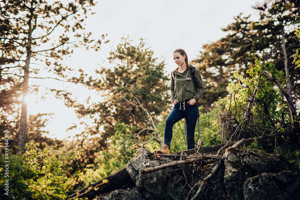 Beautiful young girl, in outdoor clothing with camelbag, standing on raised rock in middle of wilderness. In background, setting sun shines through the trees, which adds warm tones to the whole scene. Beautiful young girl, in outdoor clothing with camelbag, standing on raised rock in middle of wilderness. In background, setting sun shines through the trees, which adds warm tones to the whole scene.