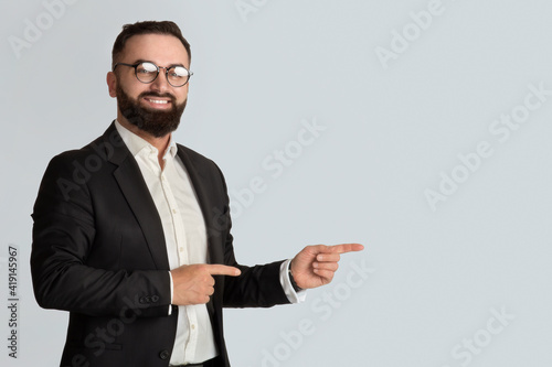 Serious businessman in office wear tand glasses smiling and pointing aside on light grey background, space for you ad photo